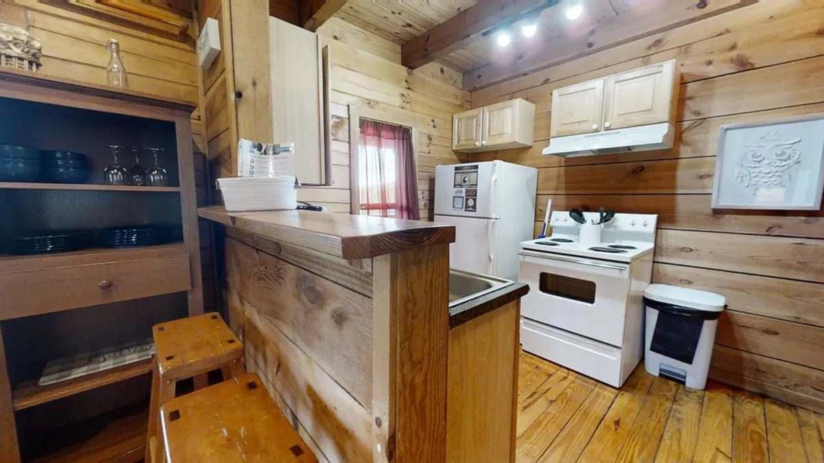 A spacious, rustic kitchen with wooden cabinets, kitchen appliances, and a row of bar stools. Natural light pours in through a window with red curtains, creating a warm, welcoming setting for guests to gather. The kitchen's open layout suggests room for group cooking, casual meals, and lively conversation.