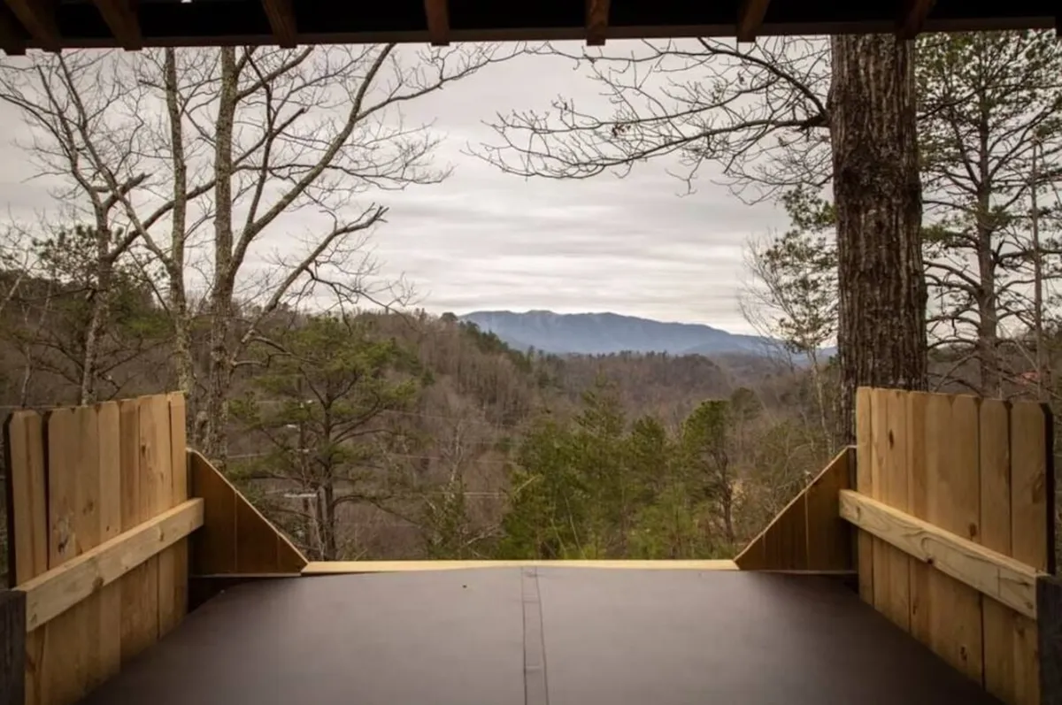 A wide-angle view showcases a panoramic mountain range with a wooden deck and surrounding trees in the foreground, giving a sense of adventure and proximity to the Smoky Mountains.