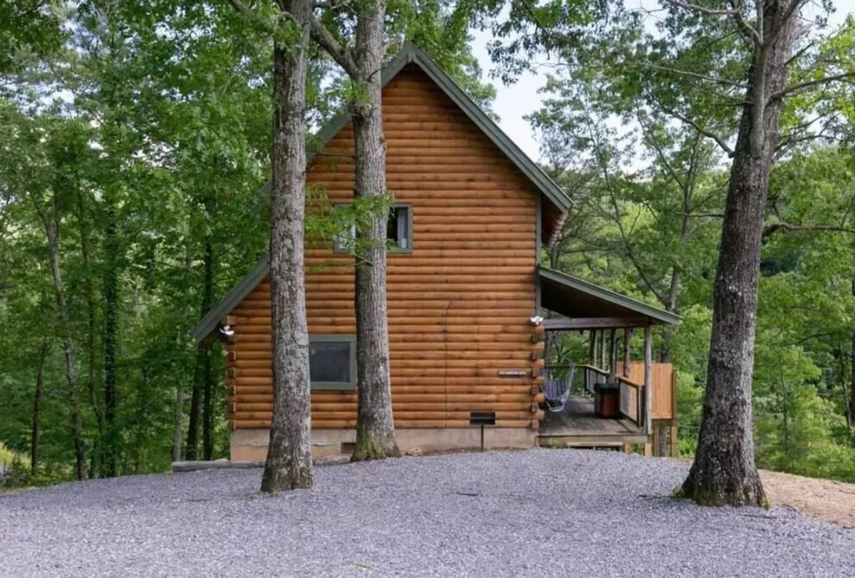 This image shows the exterior of a log cabin nestled among trees, with a level gravel driveway and a welcoming front porch. The setting is tranquil, and the driveway appears suitable for parking two cars, supporting the convenience narrative.