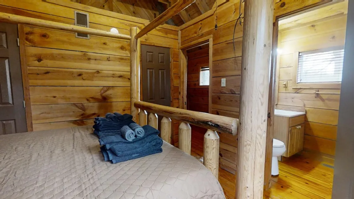 The image displays a rustic bedroom with a log bed frame, towels neatly placed on the bed, and natural wooden walls, evoking a sense of comfort and retreat. The setting suggests a peaceful environment, ideal for unwinding after a day of exploration.
