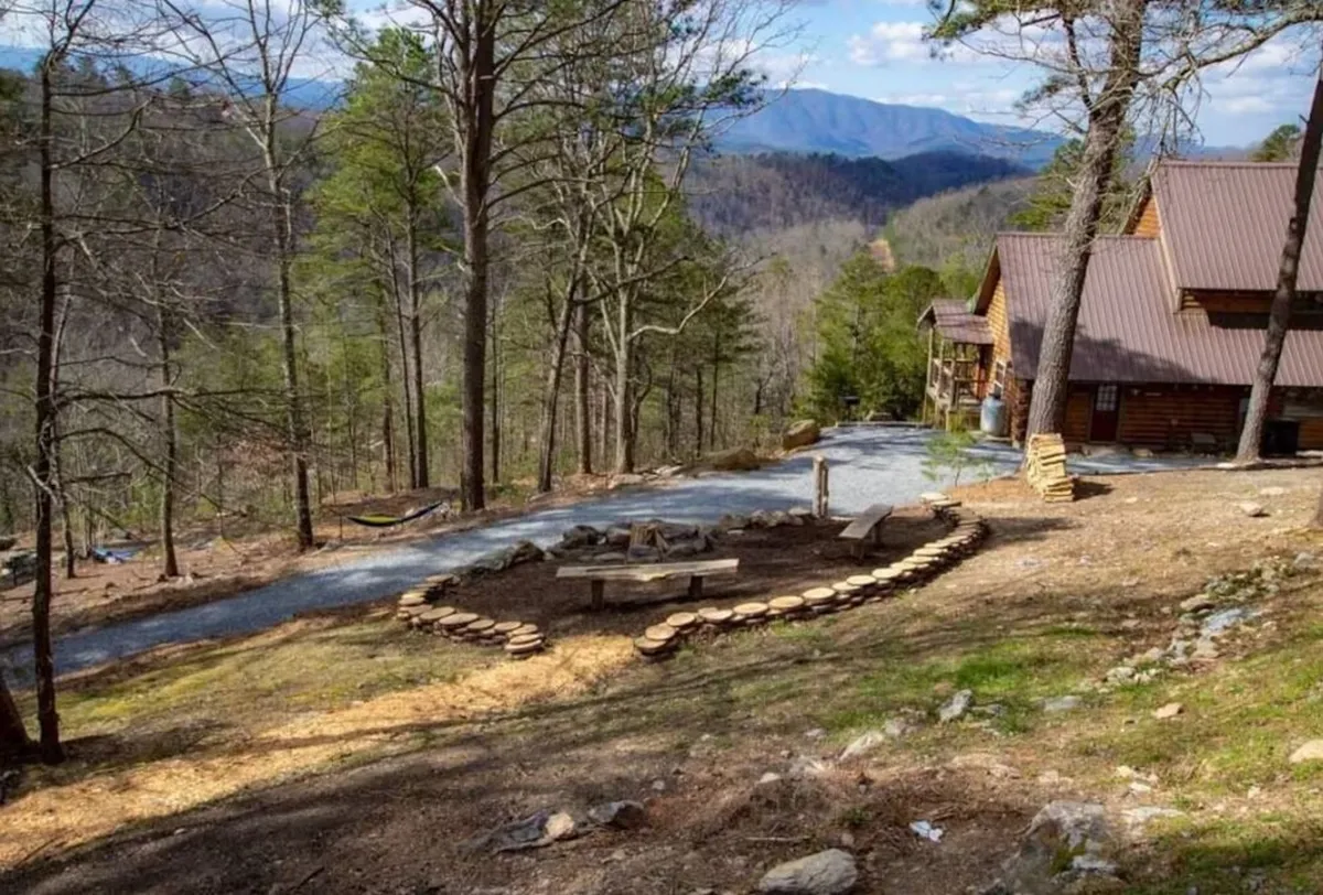 Depicts a cozy outdoor fire pit area with log seating, set against the backdrop of mountains and the cabin. The scene suggests evening gatherings, storytelling, and unwinding beneath the stars—perfect for family bonding after active days.