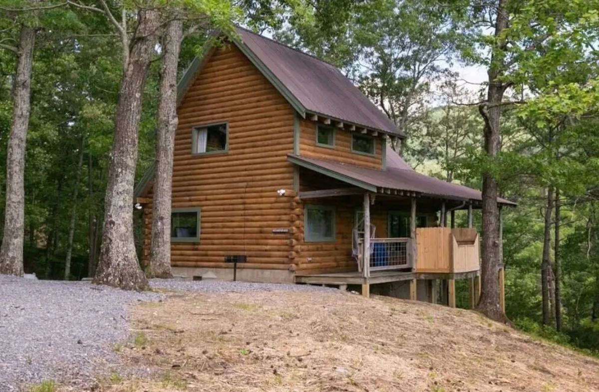 A tranquil exterior view of the log cabin nestled among trees, featuring an inviting deck area perfect for gatherings or quiet moments. The image highlights the cabin’s harmonious integration with nature and suggests the spaciousness and versatility of the outdoor space.