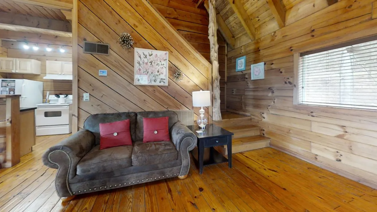 A comfortable living area featuring a sofa with red cushions, a wooden staircase leading to the upstairs bedroom, decorative wall art, and a table lamp. The room feels welcoming, perfect for socializing or relaxing, and includes a sofa that doubles as a sleeper to accommodate extra guests.