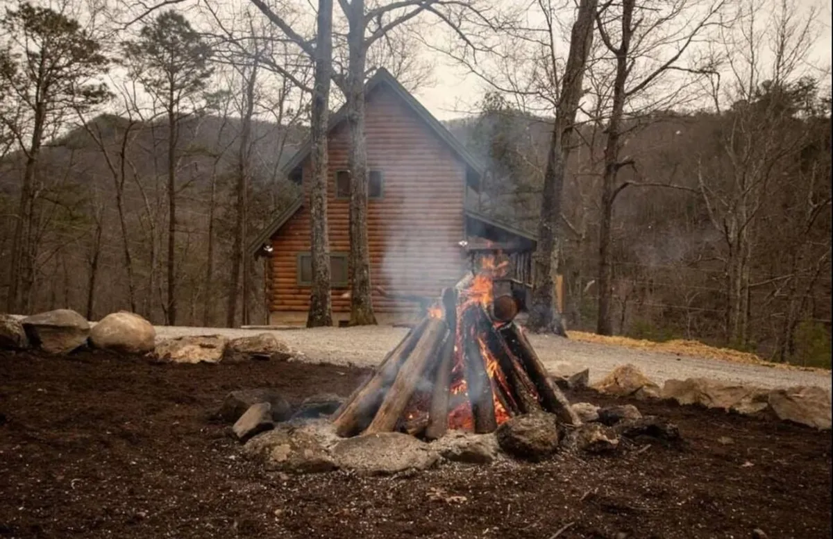 A glowing campfire surrounded by people gathered near a rustic cabin, with surrounding trees creating a cozy, communal atmosphere. The fire pit area is the central point, with marshmallows roasting and the warm light suggesting evening gatherings and shared stories.