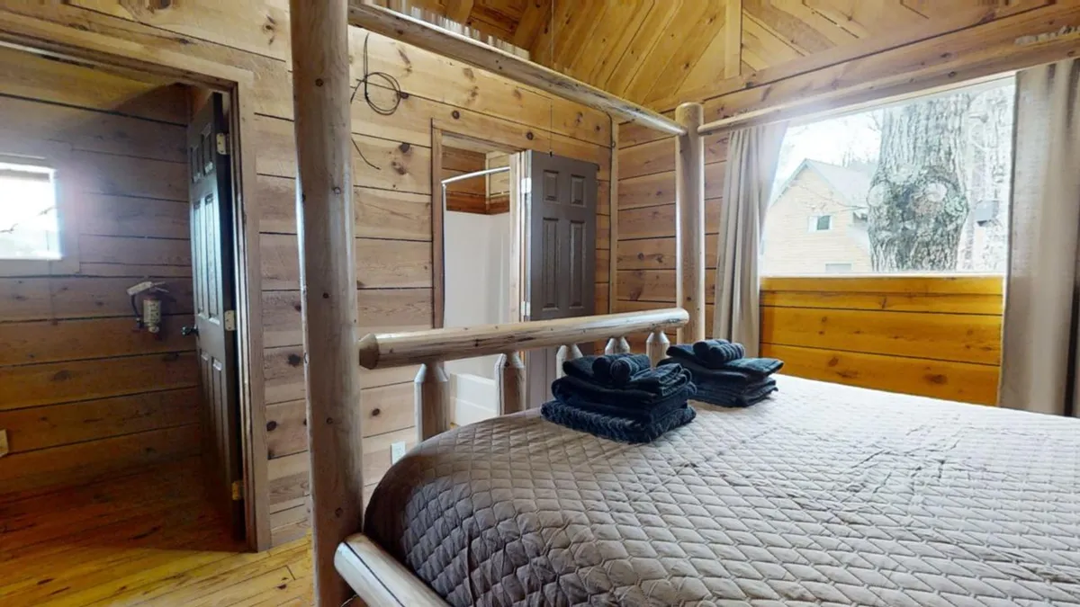 This image features a cozy wooden bed with plaid bedding, towels artfully arranged, and a window offering a view of trees just outside. The soft, natural light coming through the window creates a warm and inviting ambiance, making it easy to imagine waking up to the sights and sounds of the forest.