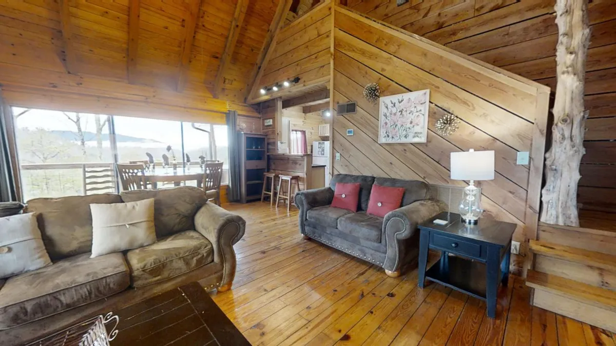 This image captures a cozy living area with a sofa adorned with decorative pillows, a wooden dining table perfect for intimate meals, and large windows offering sunset mountain views. Warm lighting and rustic decor create a welcoming, romantic atmosphere.