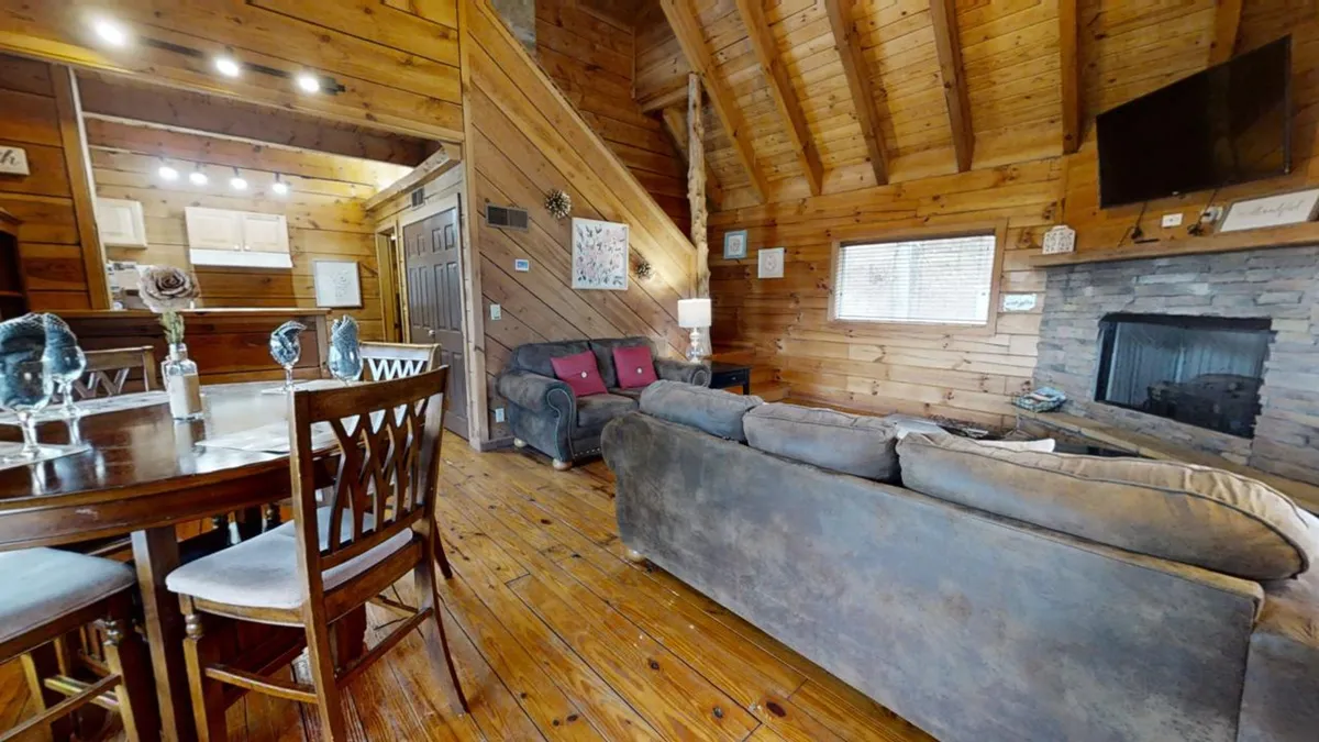The image features an open-plan living and dining area with exposed wooden beams, a striking stone fireplace, a spacious dining table surrounded by chairs, and plush sofas arranged for both comfort and conversation. Large windows flood the room with natural light, contributing to the warm and friendly atmosphere.