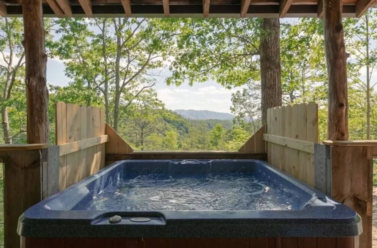 Shows a steaming hot tub on the cabin’s deck, surrounded by trees and offering a panoramic mountain view. The tranquil setting is ideal for soothing tired muscles after hikes and soaking in the serene natural beauty.