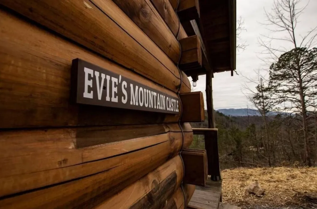 This image features the log cabin structure of Evie's Mountain Castle, prominently displaying its sign and surrounded by lush trees and distant mountains. The setting exudes a sense of adventure and immersion in nature, with the inviting porch suggesting relaxation after a day spent exploring the outdoors.